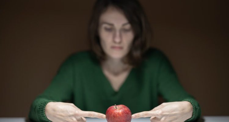 Blurred woman in green sweater points at a focused red apple on a table indoors.
